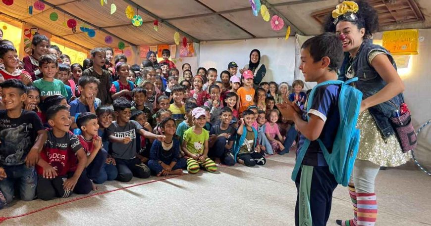 A boy with a backpack stands next to a clown in front of a crowd of children.