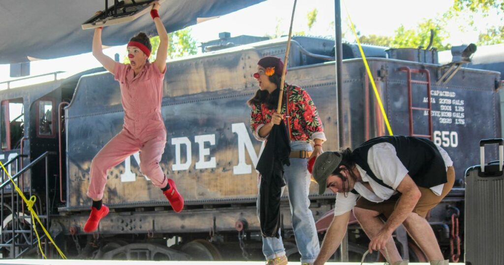 Three clowns on stage, one of them is jumping in the air with a table abover her head.