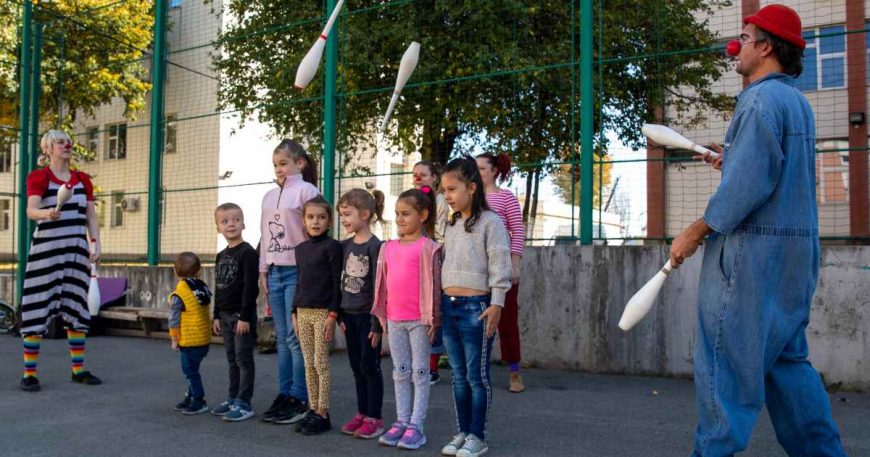 Pair of clowns juggling pins in front of a group of kids