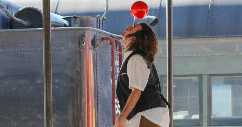 A clown bounces a ball on his head in front of a train