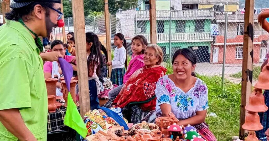 A clown and a smiling woman at a table where she is selling clay crafts.