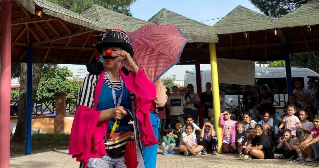 A woman clown with an umbrella wears a dark mustache.