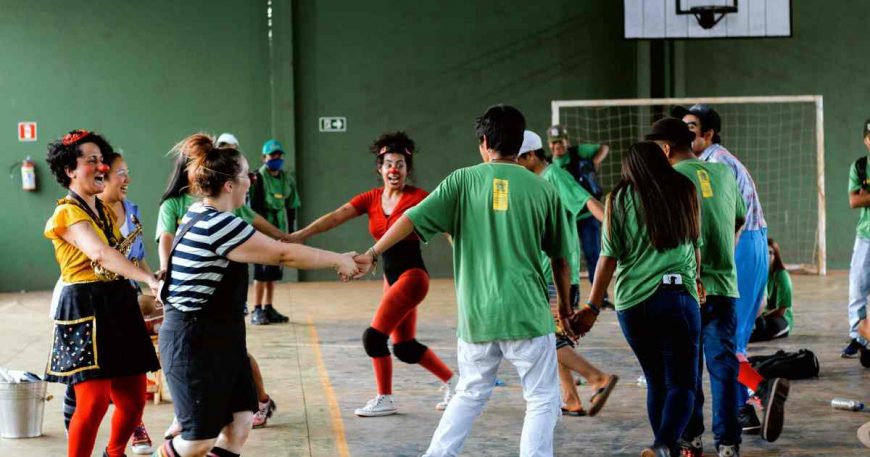 Clowns and adolescents hold hands and run in a circle in a ball court.