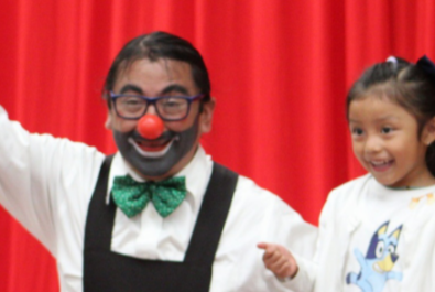 A clown kneeling next to small child with a red curtain backdrop.